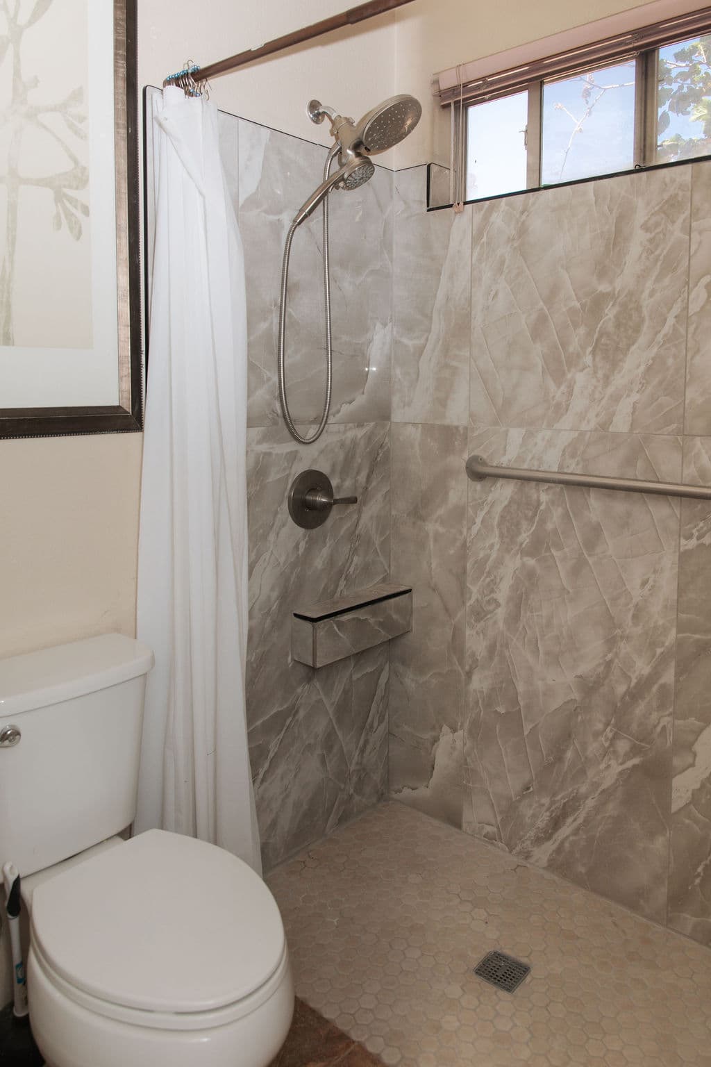 A bathroom with a curbless, walk-in tiled shower, a handheld shower head, a built-in bench ledge and grab bars.