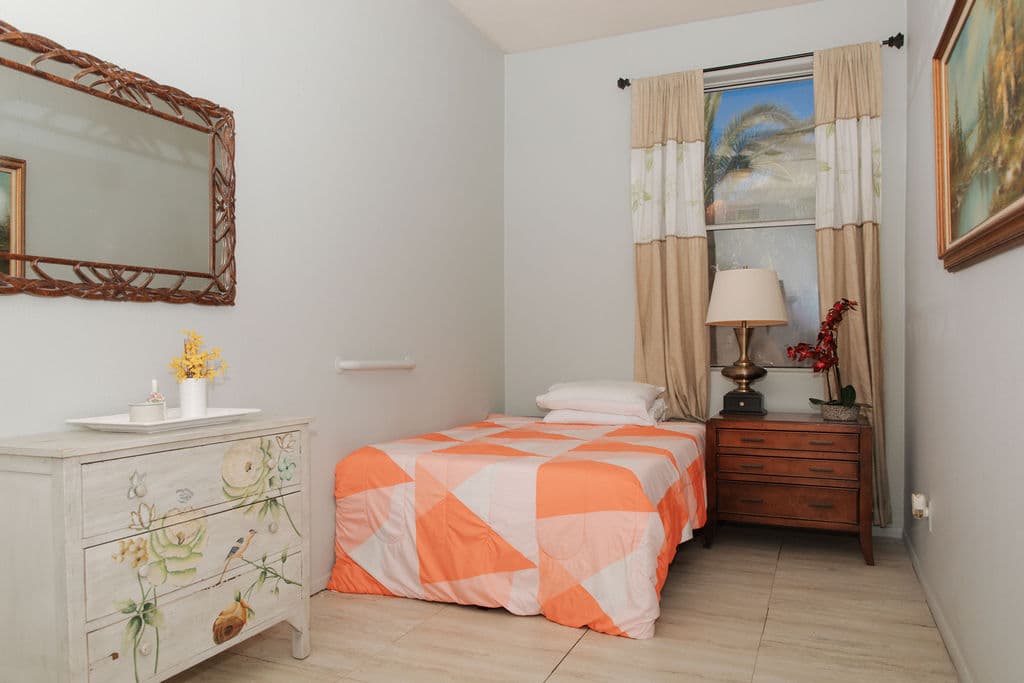A bedroom with a hand-painted floral dresser, a single bed with a peach quilt and a window framed by patterned curtains.