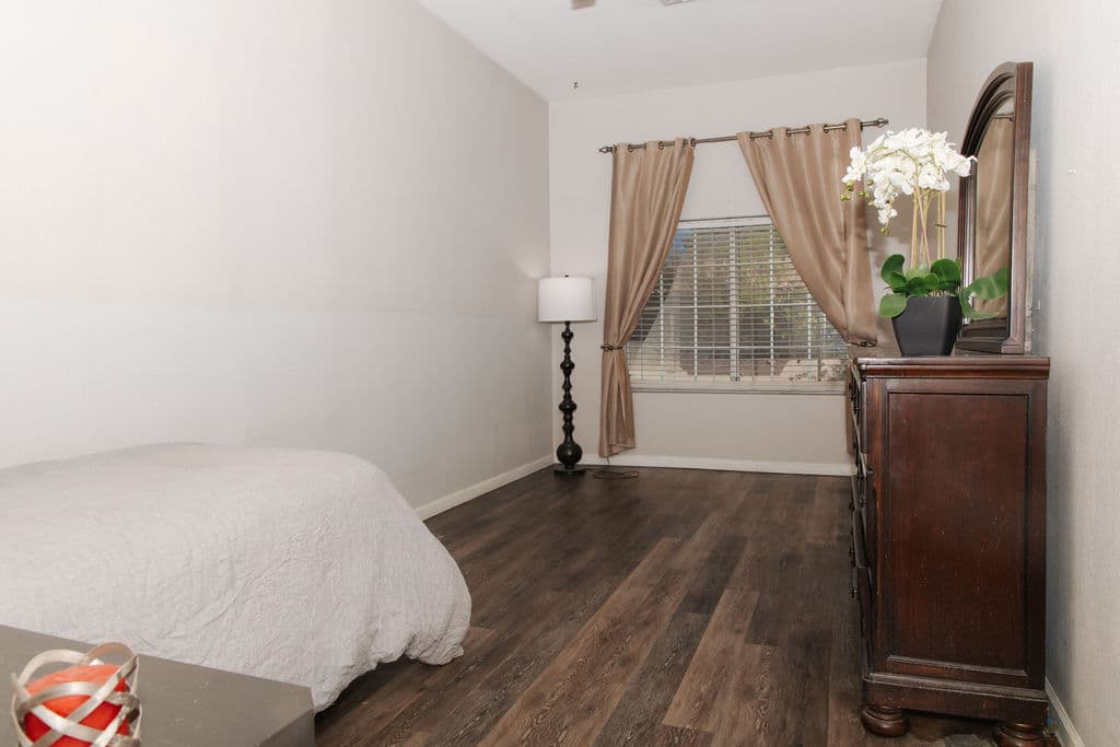 A calm bedroom with a made bed, a dark wood dresser topped with a white orchid, soft curtains and wood floors.