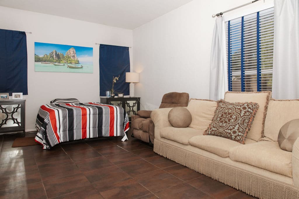 A spacious private room with a bed, a cream sofa with cushions, a recliner and wood flooring, lit by a tall window.