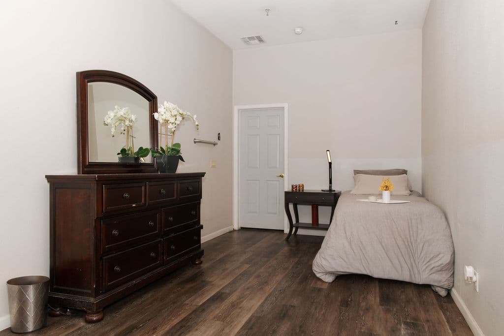 A private room with a single bed, a bedside table and a dark wood dresser with a mirror and orchids.