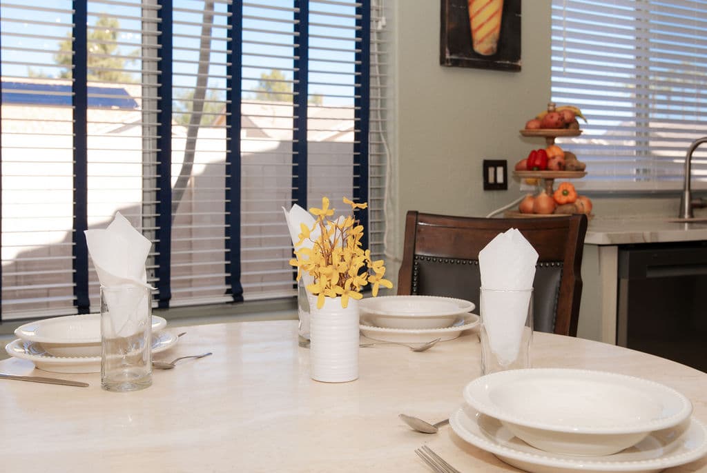 A close detail of a set table: white plates, folded napkins in glasses and a small vase of yellow flowers by the window.